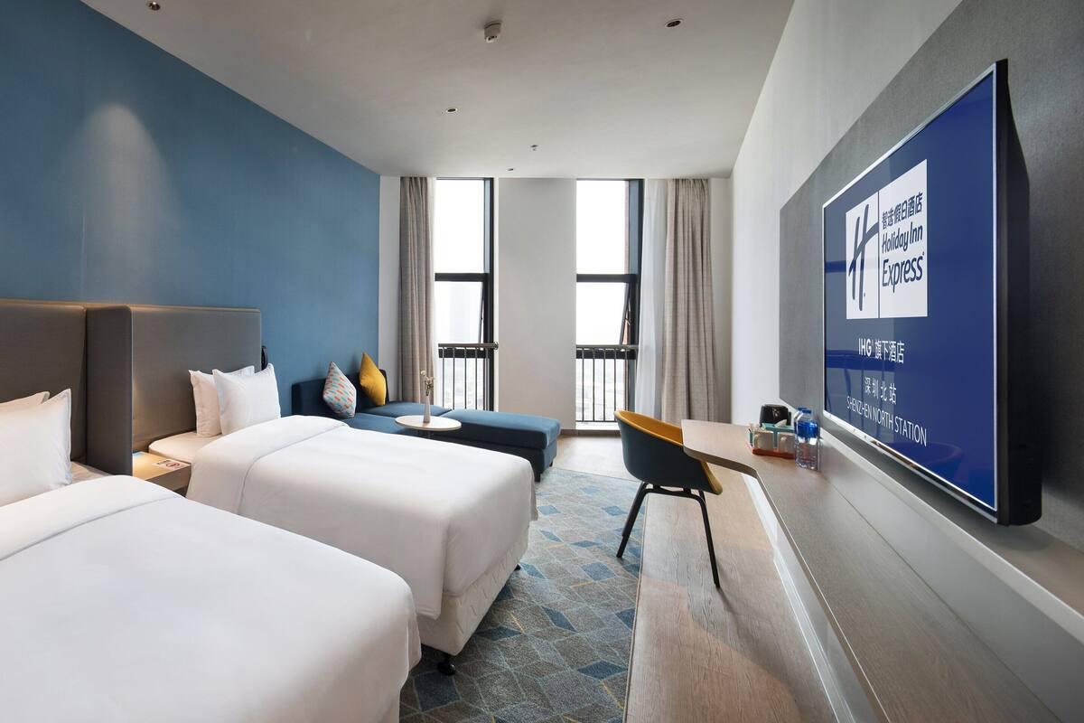 A hotel room with two beds, a desk, a chair, and a large TV screen displaying the Holiday Inn Express logo.