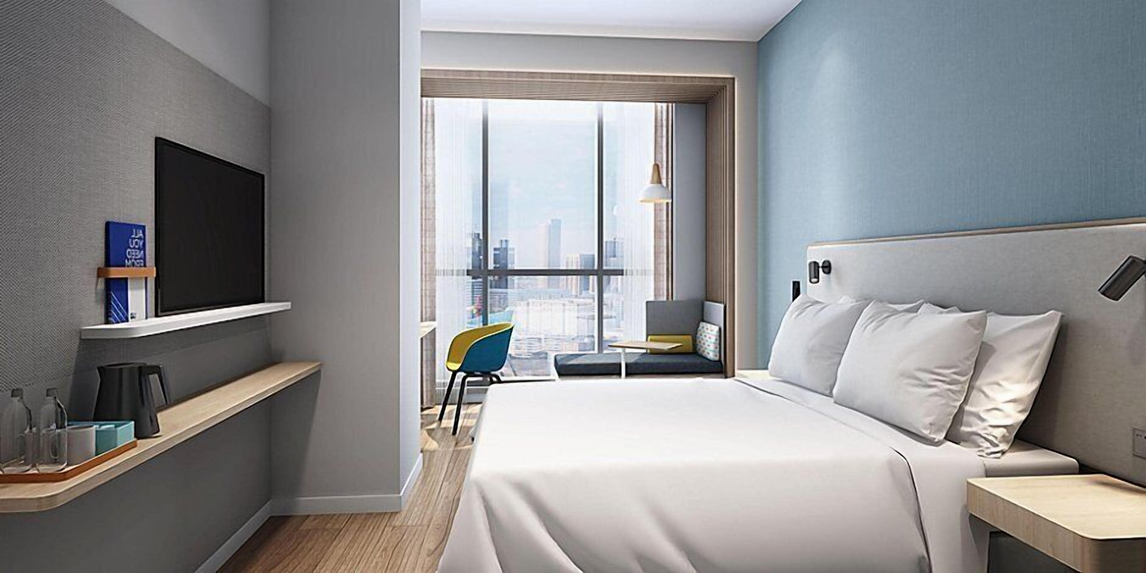 A modern hotel room with a large bed, a television mounted on the wall, a small desk, and a window with a city view.