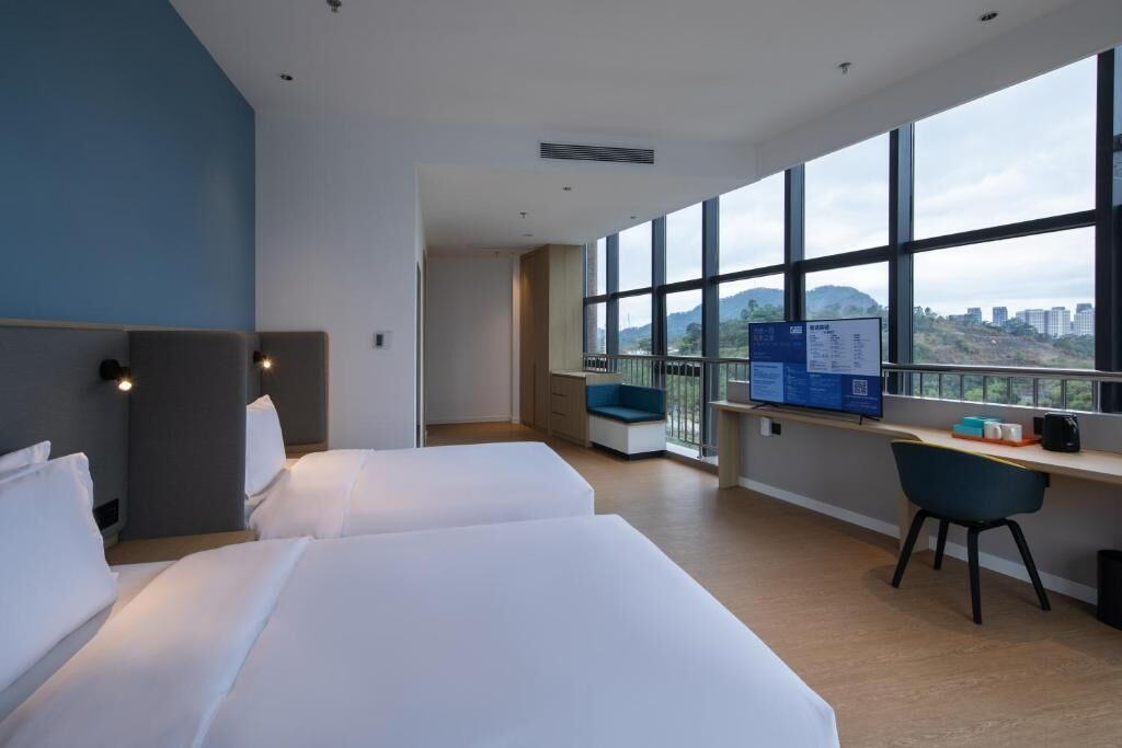 A hotel room with two beds, a desk, a chair, and a large window offering a city view.