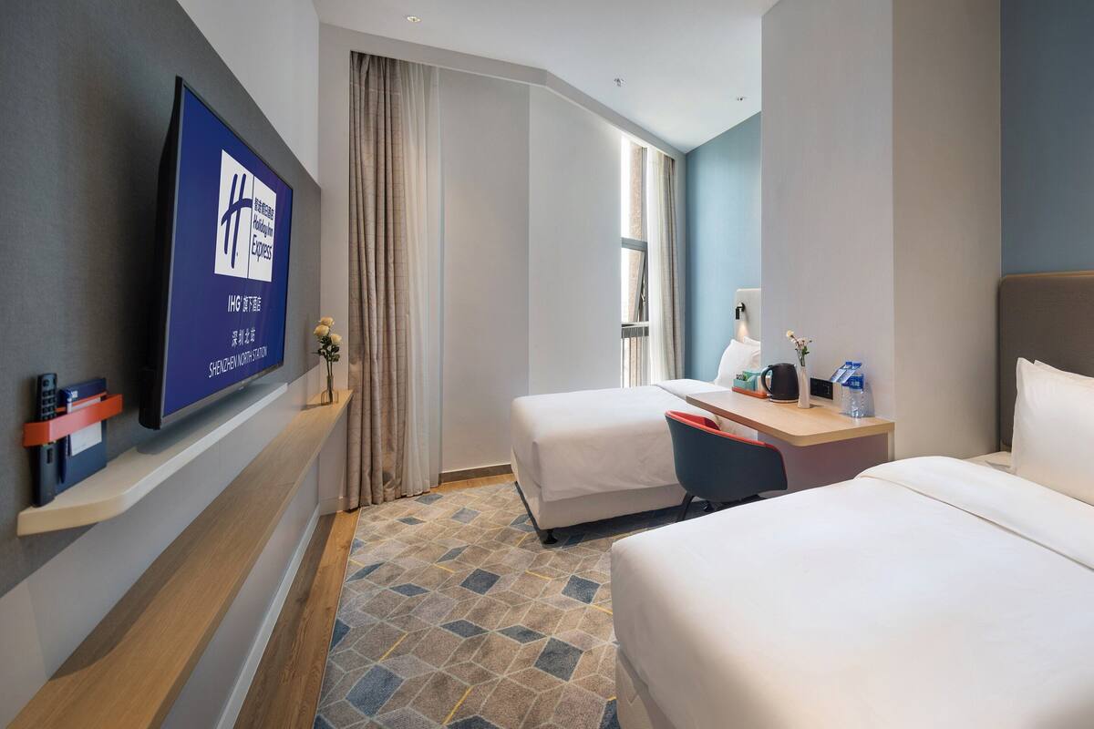 A hotel room with a large bed, a smaller bed, a desk with a chair, a TV mounted on the wall, and a window with curtains.