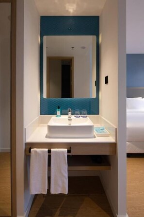 Een moderne badkamer met een witte wasbak, een muur betegeld met blauwe tegels en een grote spiegel. Er hangen twee witte handdoeken aan een rek onder de wasbak.