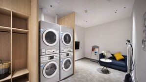A laundry room with four stacked front-loading washing machines, a small table with a vase, and a blue sofa with yellow cushions. - Holiday Inn Express Shenzhen North Station by IHG (Shenzhen)