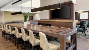Un espace repas moderne dans un hôtel, avec une longue table en bois, des chaises à revêtement beige, un grand téléviseur à écran plat et une plante en pot.