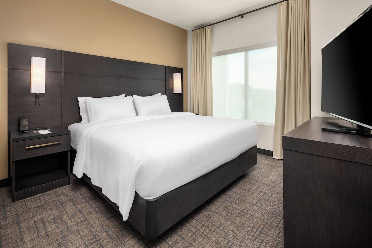 A hotel room with a large bed, a nightstand with a lamp, a desk with a flat-screen TV, and a window with curtains.