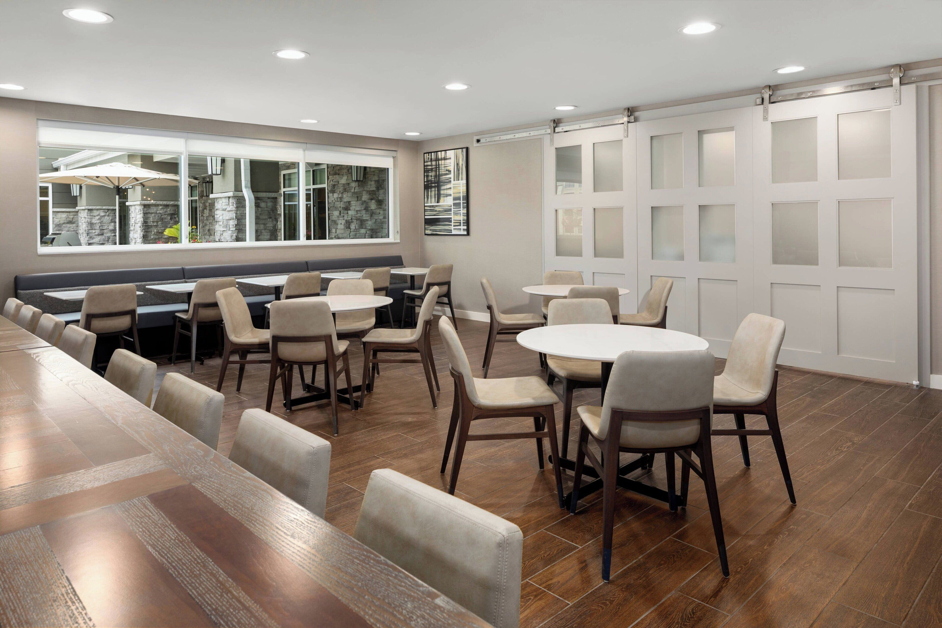 Un comedor moderno con una larga mesa de madera y varias mesas redondas, rodeado de sillas.