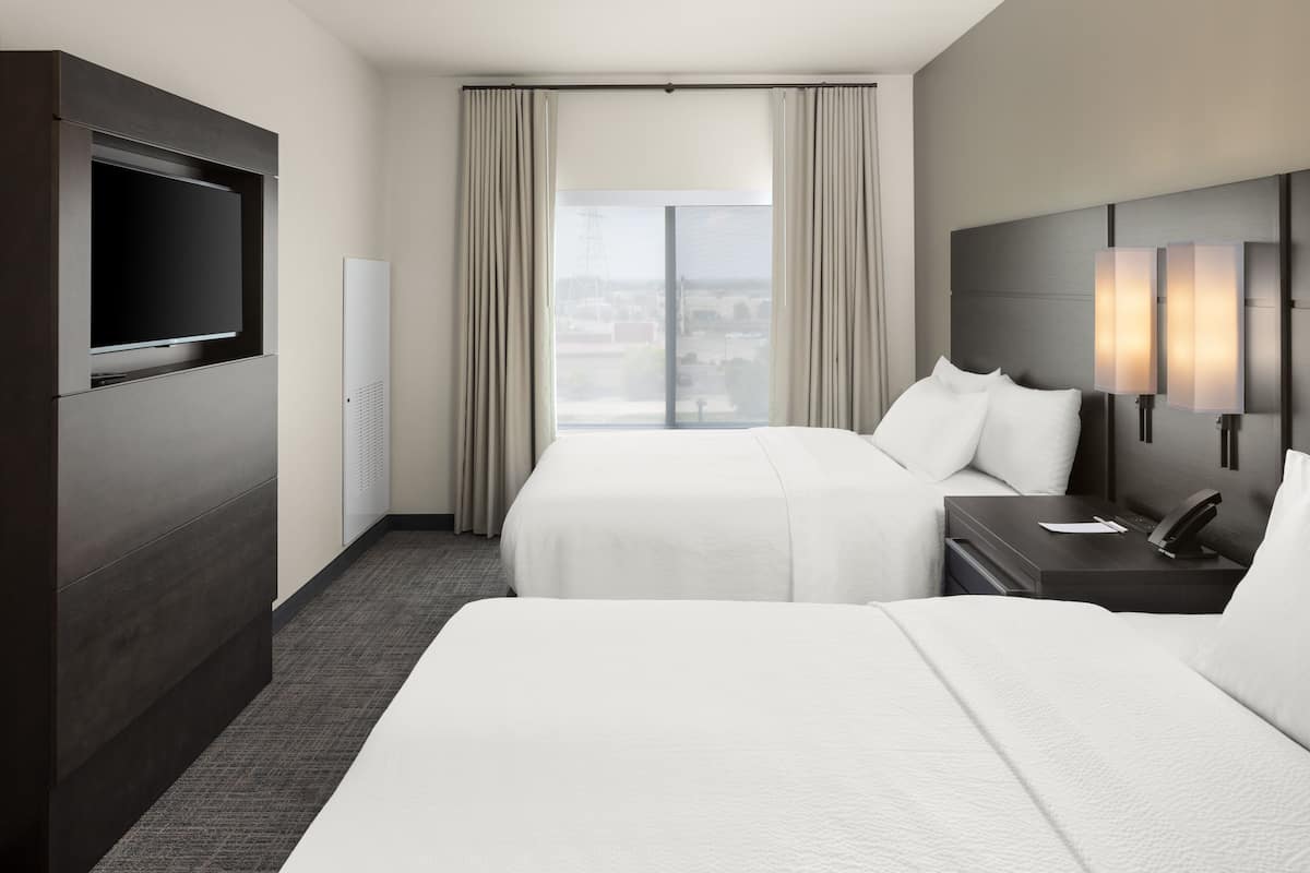 A hotel room with two beds, a flat-screen TV, a bedside table with a lamp, and a large window with curtains.