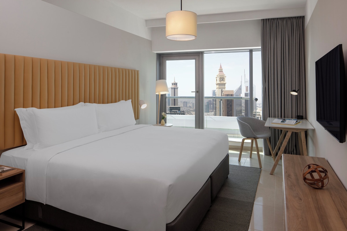 A modern hotel room with a large bed, a desk, and a view of a cityscape.