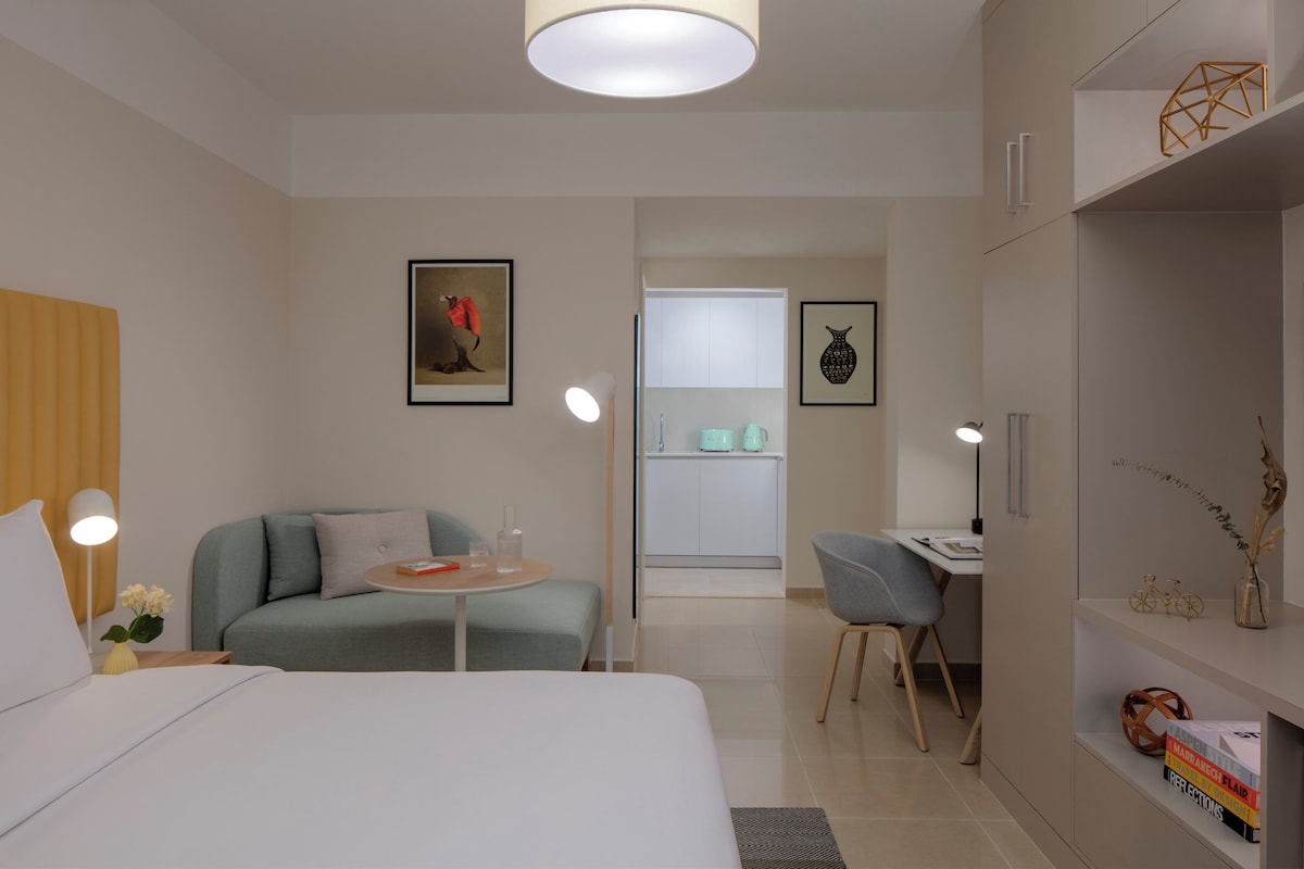 A modern hotel room with a bed, a seating area with a table, a desk, and a kitchenette.