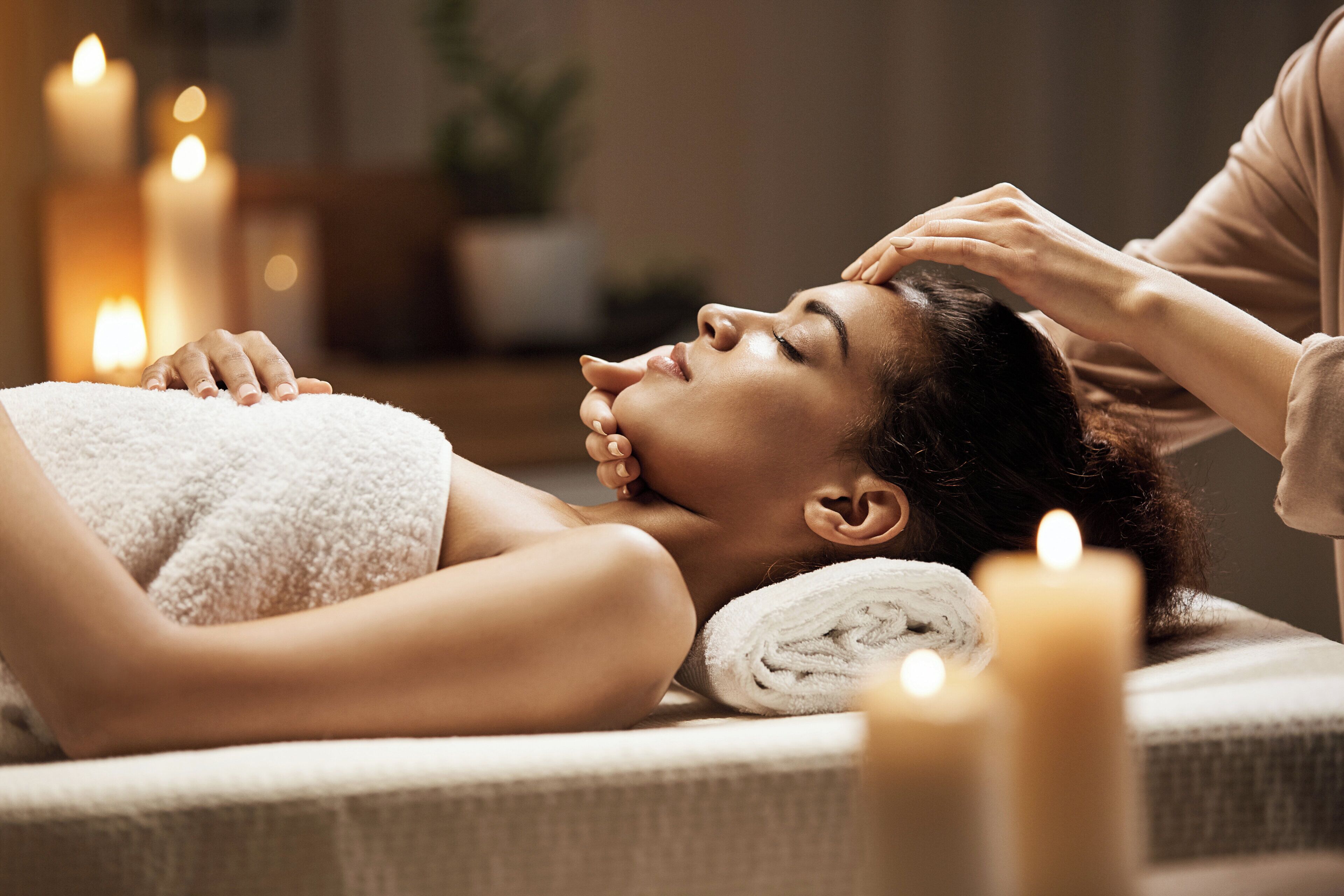 Eine Person liegt auf einer Massageliege, umhüllt von einem Handtuch, und erhält eine Kopfmassage. Kerzen erzeugen im Raum eine entspannte, warme Atmosphäre.