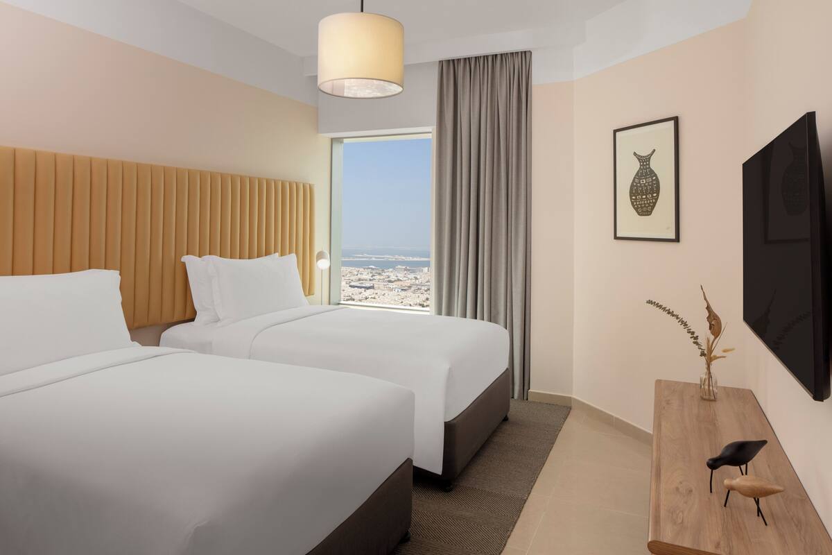 Une chambre d’hôtel avec deux lits, une grande fenêtre donnant sur un beau paysage, un bureau en bois et un téléviseur fixé au mur.