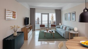 Una sala moderna con sofá, mesa de centro y mesa de comedor. Hay un televisor sobre un mueble de madera, y vistas de la ciudad a través de amplias ventanas.
