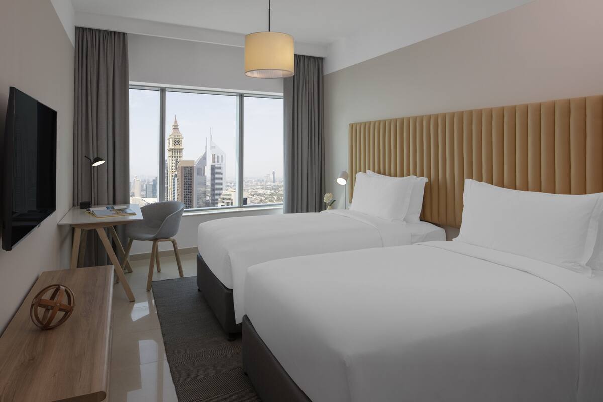 Une chambre d’hôtel avec deux lits, un bureau, une chaise, une télévision et une grande fenêtre donnant sur la ville.
