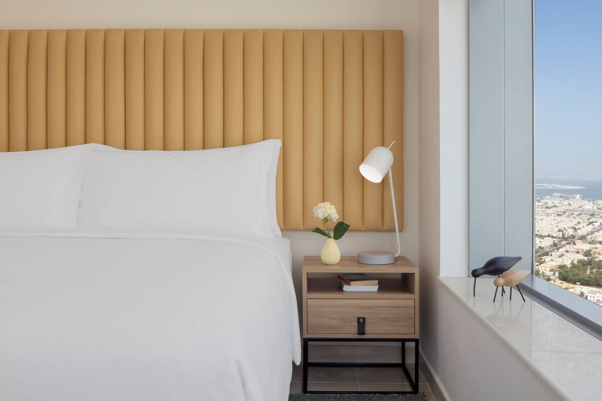 Une chambre d’hôtel moderne avec un grand lit, une table de chevet avec une lampe, un vase rempli de fleurs et une vue sur la ville par une grande fenêtre.