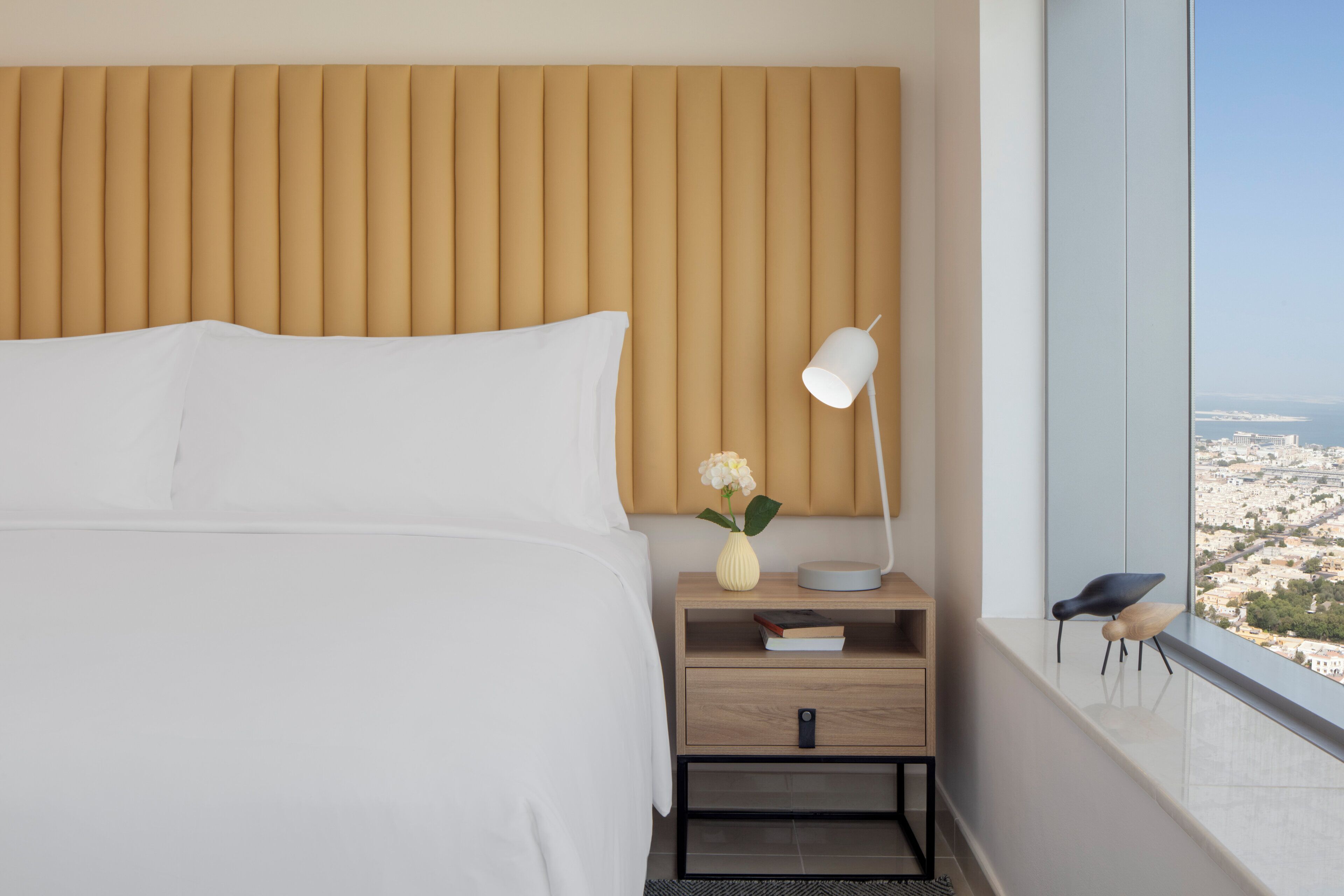 A hotel room with a bed, bedside table, lamp, vase with flowers, and a view of a cityscape through a window.
