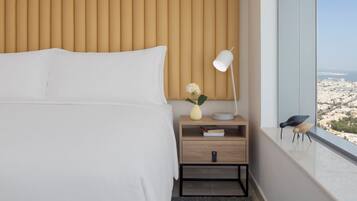 A hotel room with a bed, bedside table, lamp, vase with flowers, and a view of a cityscape through a window.