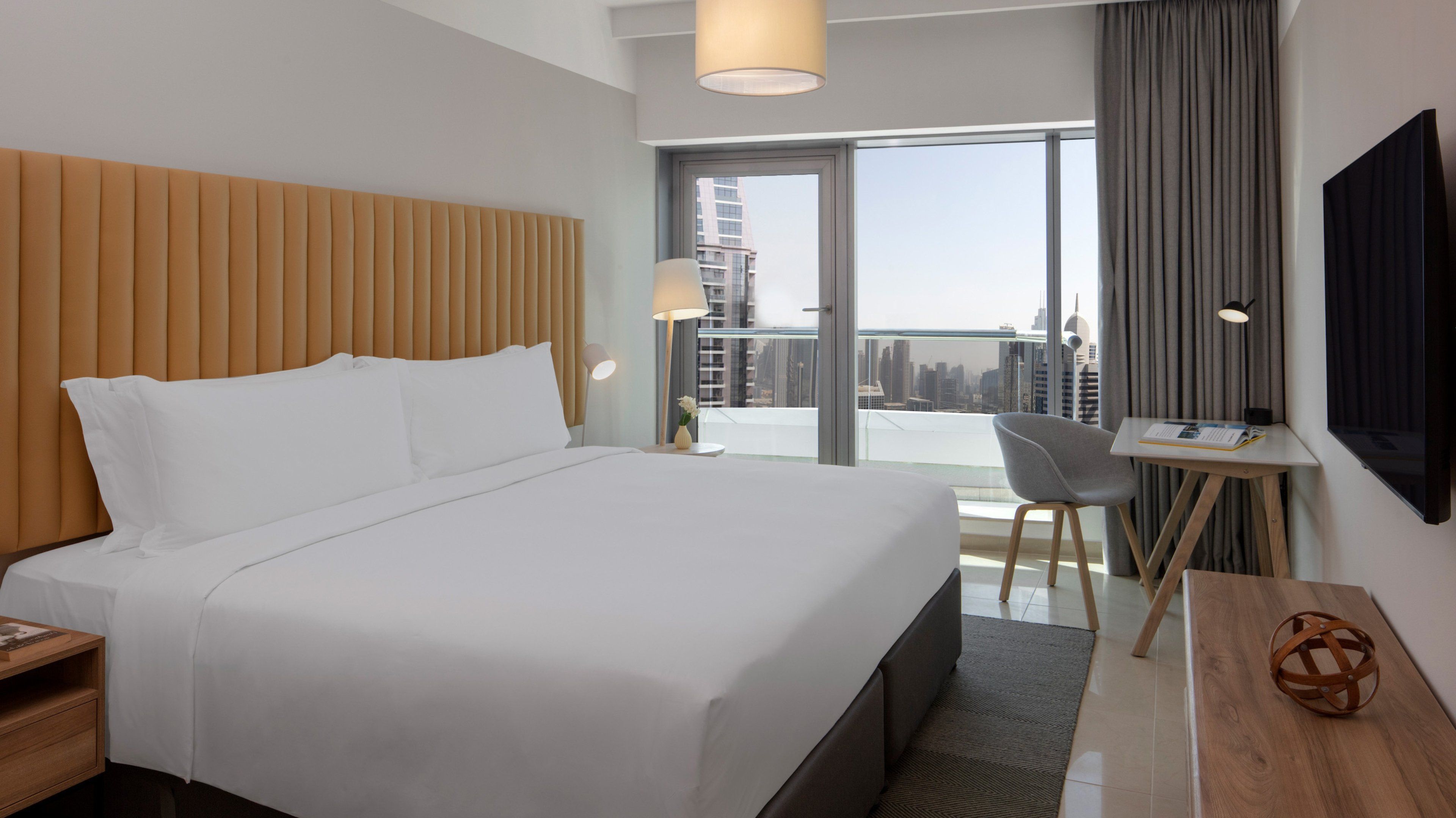 A modern hotel room with a large bed, a desk, a chair, and a view of the city skyline through the window.