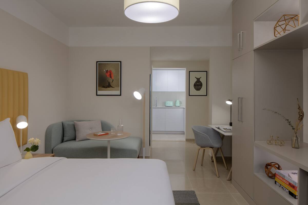 Une chambre d’hôtel moderne avec un lit, un canapé, un bureau et une chaise. On y trouve une petite table avec une lampe, ainsi que des œuvres d’art accrochées aux murs.