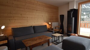 Een gezellige woonkamer met een houten wand, een bank, een salontafel, een bijzettafeltje met een lamp en een open haard.