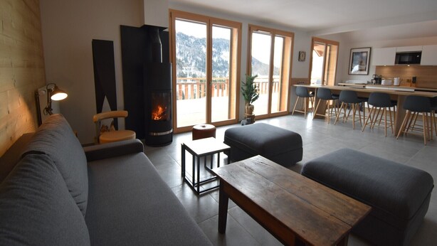 Een moderne woonkamer met een open haard, een bank, een salontafel en een eethoek met stoelen, met een keuken op de achtergrond.