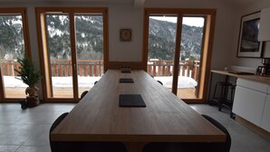 Een lange houten tafel met zwarte tafelmatten in een ruimte met grote ramen met uitzicht op een besneeuwde veranda en bergen.