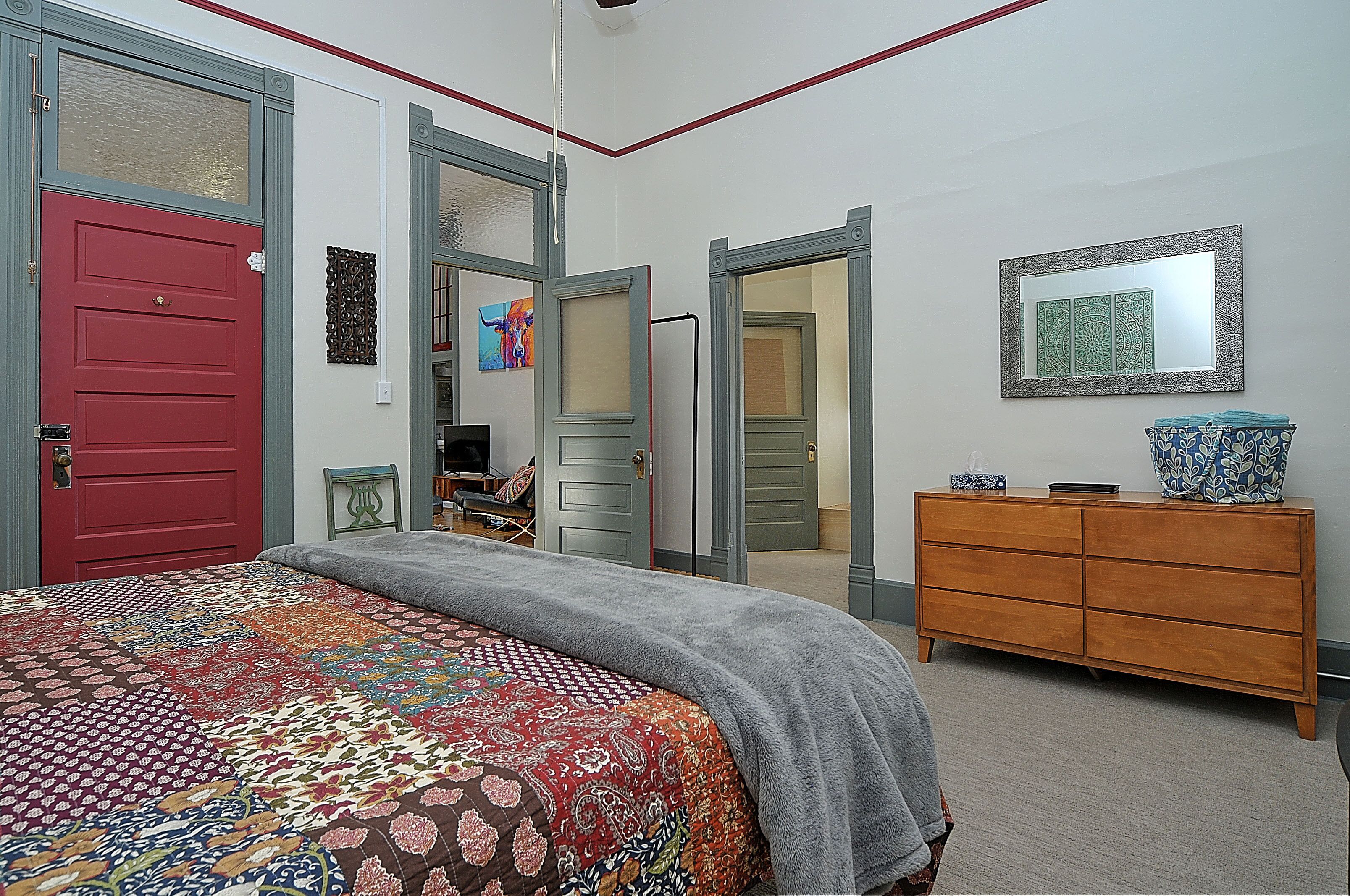 A bedroom with a red door, a gray bedspread, a wooden dresser, and a mirror on the wall.