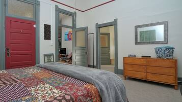 A bedroom with a red door, a gray bedspread, a wooden dresser, and a mirror on the wall.