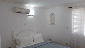 A bedroom with a bed, white walls, a window with a white frame, and an air conditioning unit mounted on the wall. - Town House with full ocean view.   (Negril)