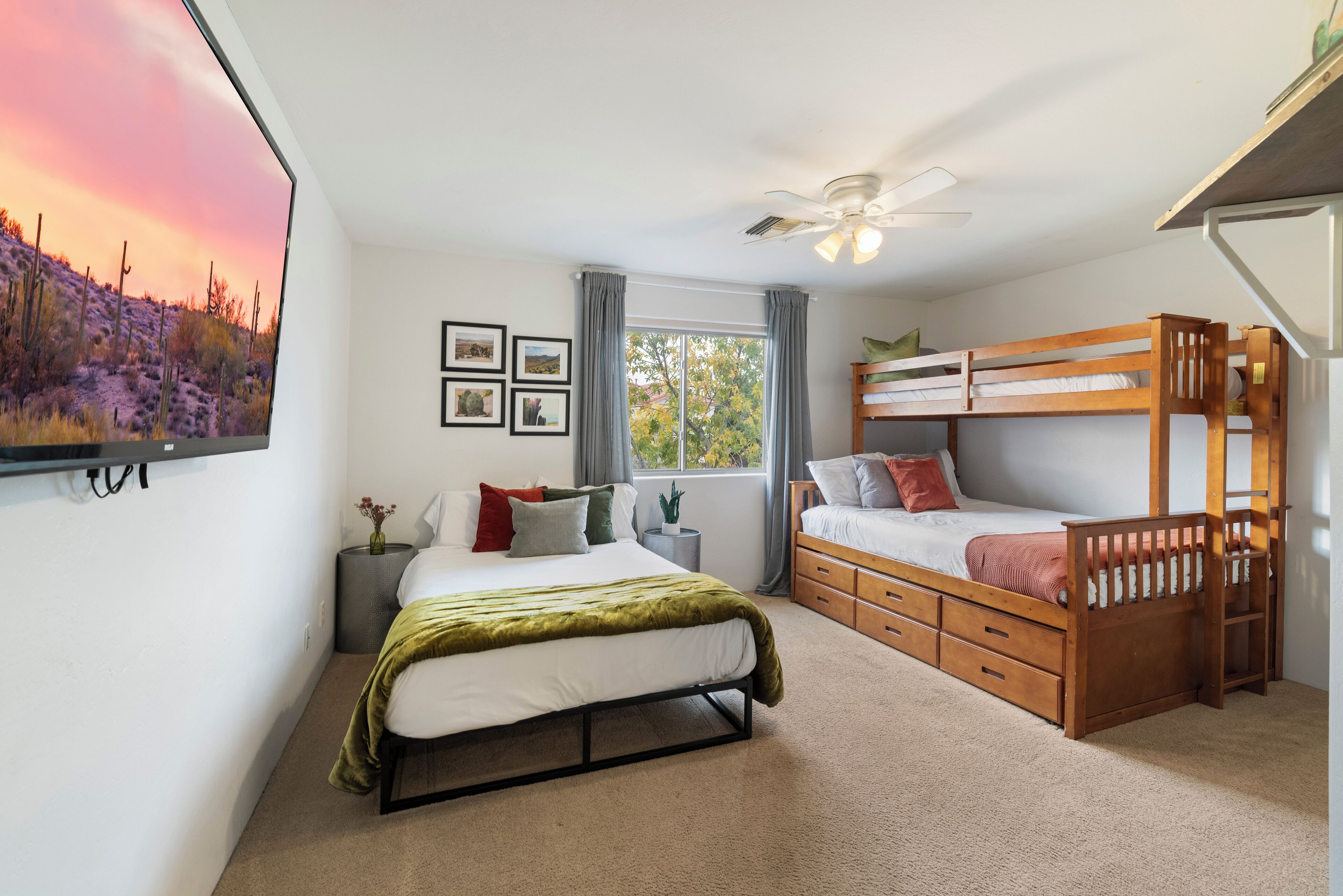 Ein Schlafzimmer mit Etagenbett, einem Einzelbett, einem Deckenventilator, einem Fenster mit Vorhängen und einem an der Wand montierten Fernseher, der eine Sonnenuntergangsszene zeigt.