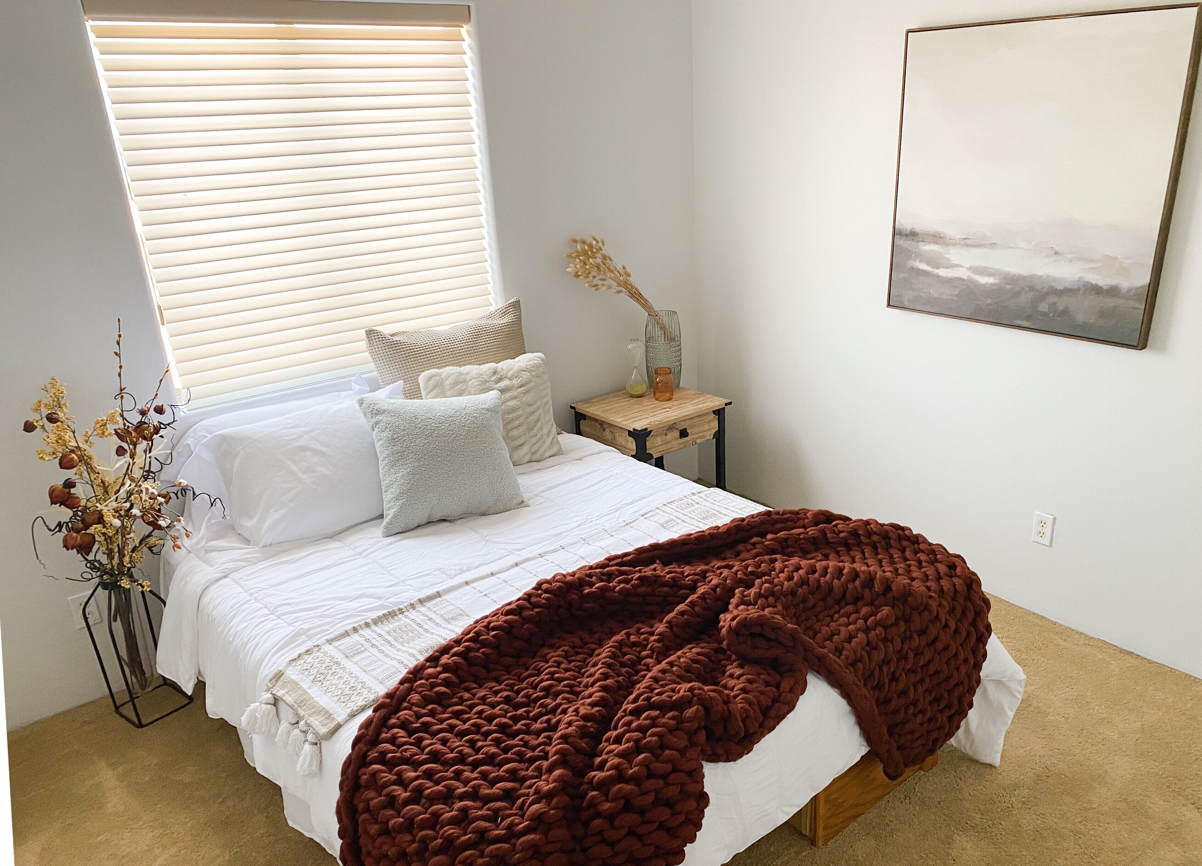 A bedroom with a bed, white bedding, a brown knitted blanket, a bedside table with a vase, and a wall-mounted painting.