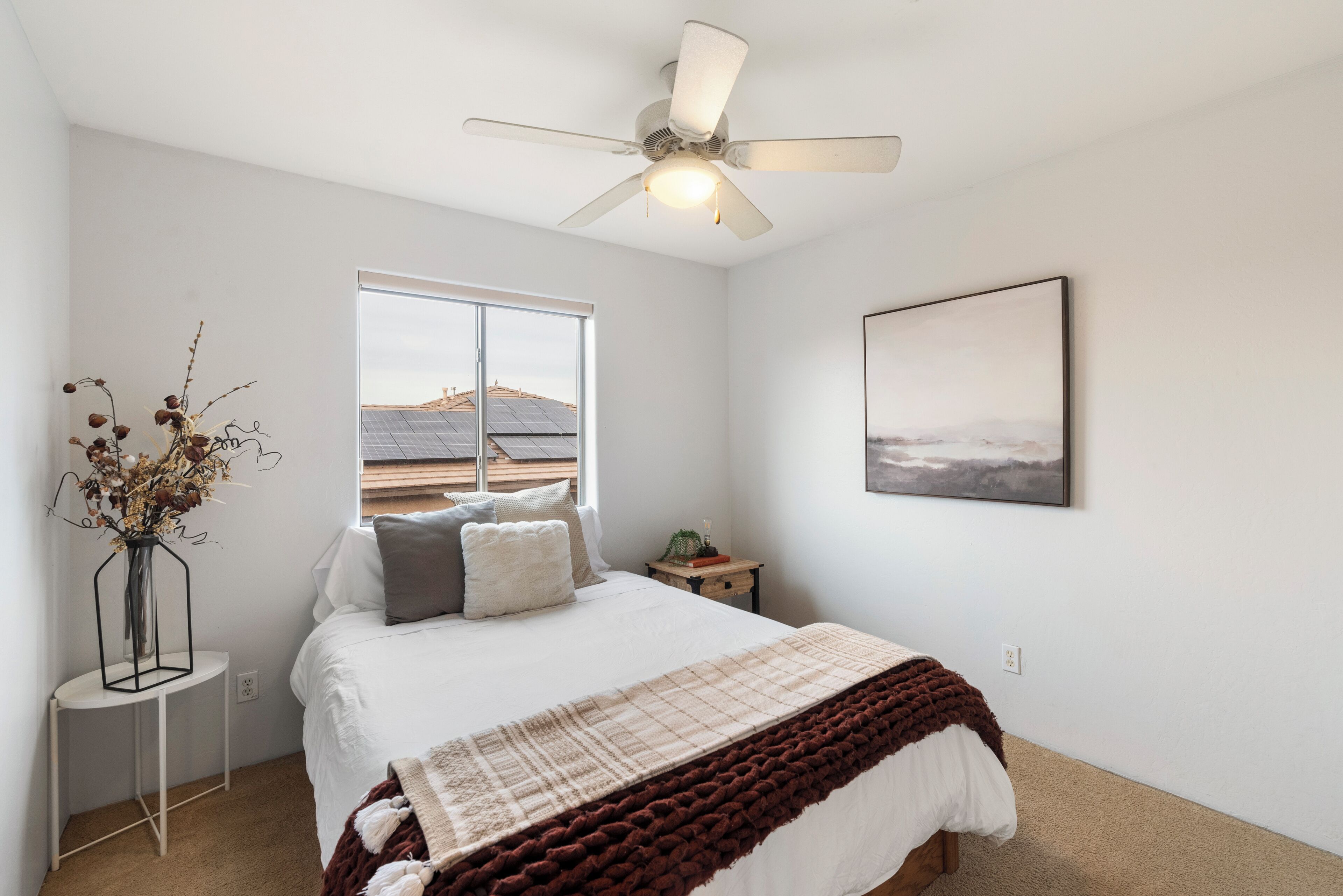 A bedroom with a bed, a window with a view of solar panels, a ceiling fan, and a framed picture on the wall.