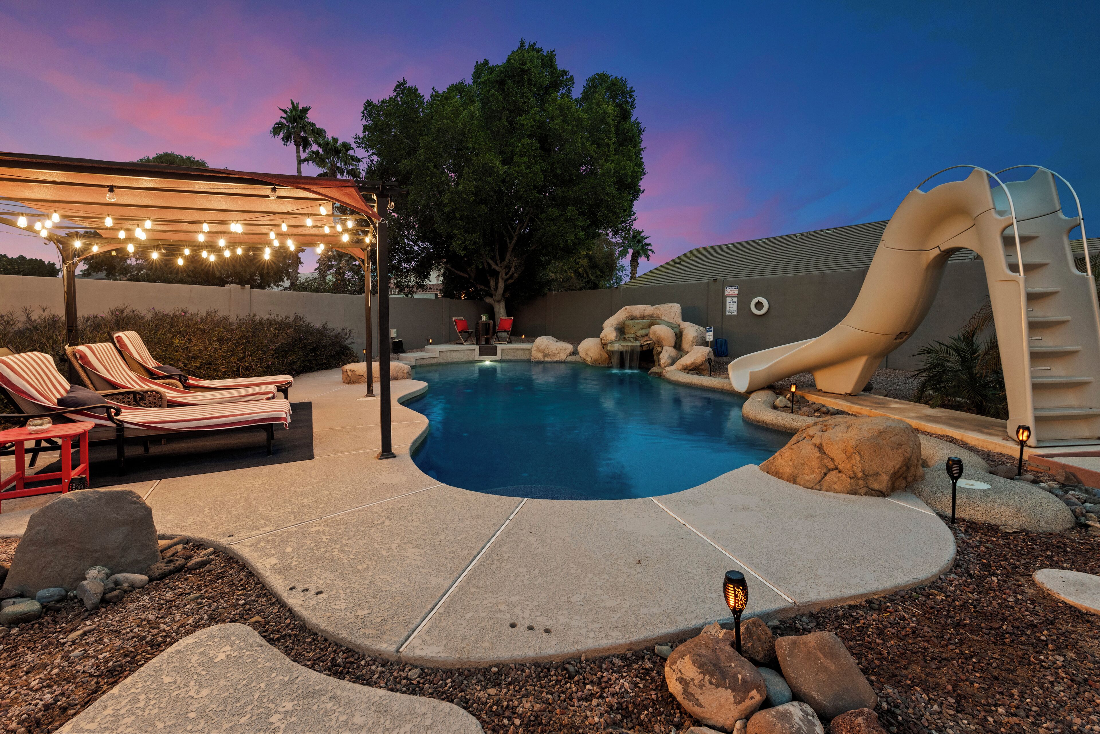 A backyard with a swimming pool, lounge chairs, a slide, and string lights.