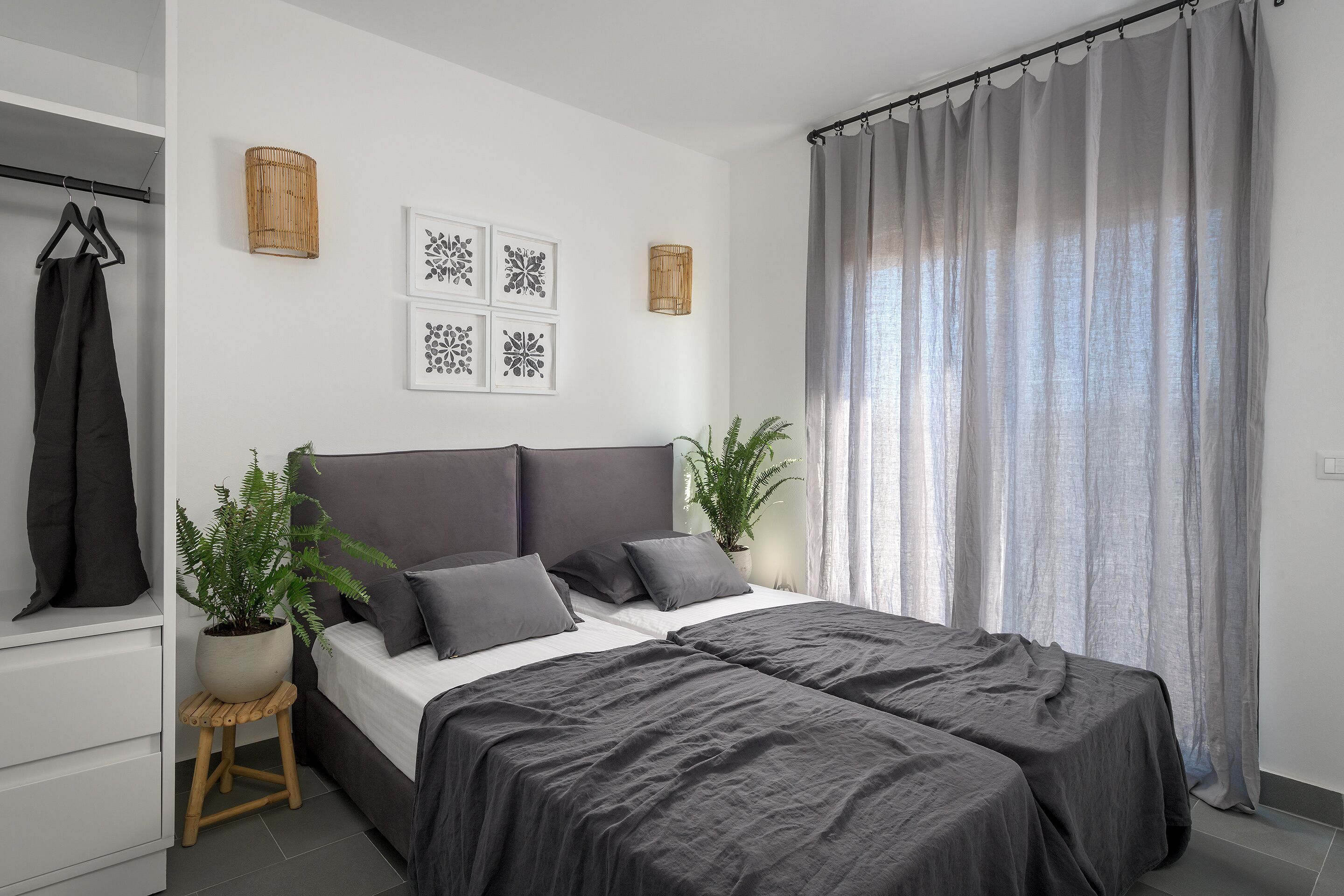 Ein modernes Schlafzimmer mit einem großen Bett, grauer Bettwäsche, einer Zimmerpflanze und Blick ins Freie durch transparente Vorhänge.