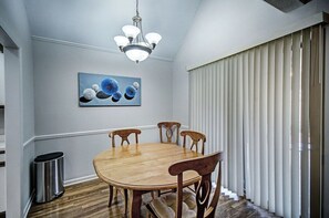 A dining room with a wooden table and chairs, a chandelier, a painting on the wall, and a window with blinds.