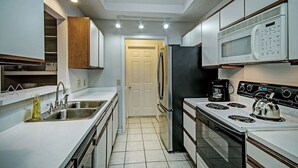 Cocina compacta con armarios blancos, pileta de acero inoxidable, refrigerador negro, microondas, estufa y cafetera.