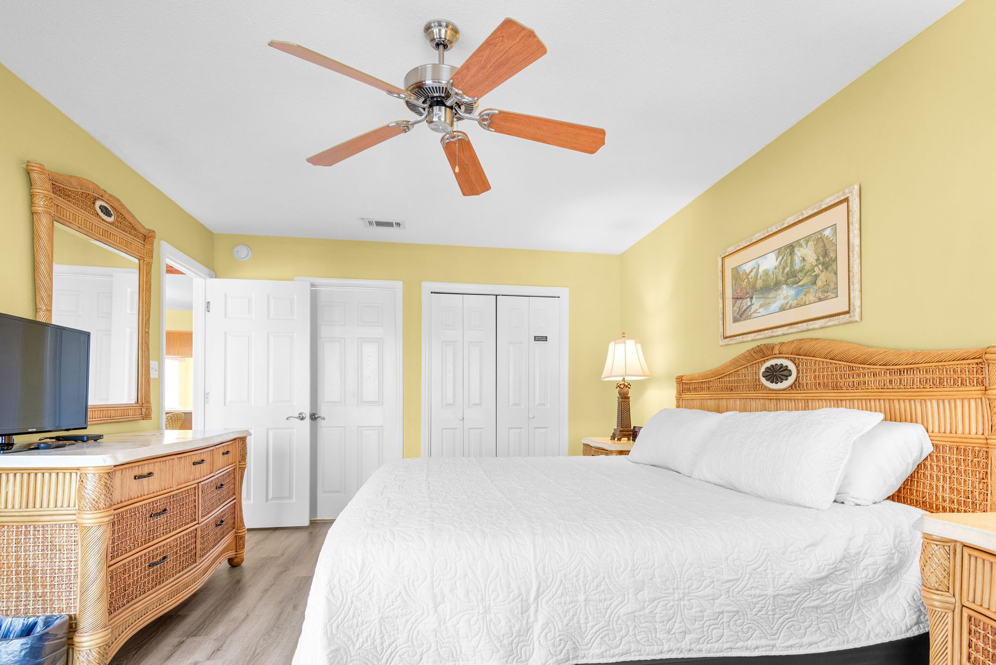 A hotel room with a bed, a television, a ceiling fan, a dresser, and a painting on the wall.
