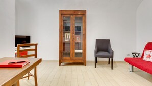 Een kamer met een houten kast, een grijze fauteuil, een rode bank, een houten tafel met een rode map, en een televisie op een standaard.