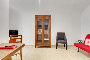 A room with a wooden cabinet, a grey armchair, a red sofa, a wooden table with a red folder, and a television on a stand.