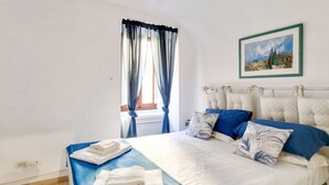 Een slaapkamer met een bed, kussens, een schilderij, een raam met gordijnen en uitzicht op het buiten.