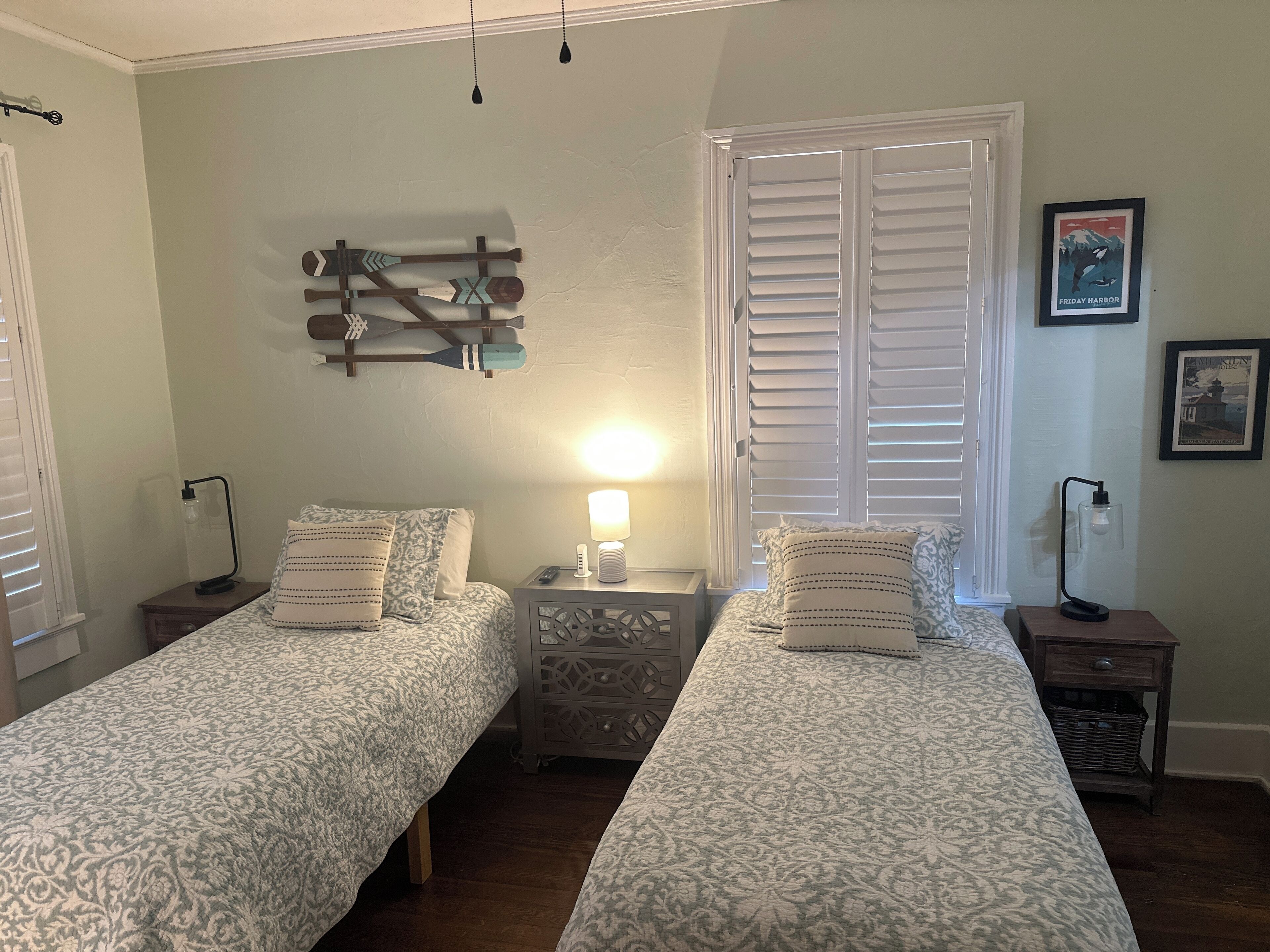 A bedroom with two beds, a nightstand with a lamp, a wall-mounted light, and a framed picture.