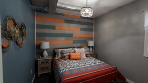 Quarto com colcha estampada, mesa de cabeceira com abajur e parede com padrão listrado.