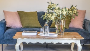 A blue sofa with green and pink cushions, a white table with a book and two glasses, and a vase with flowers. - Seaside Retreat - Premium Luxe Accommodation: 3rd night complimentary: winter 26 (Port Elliot)