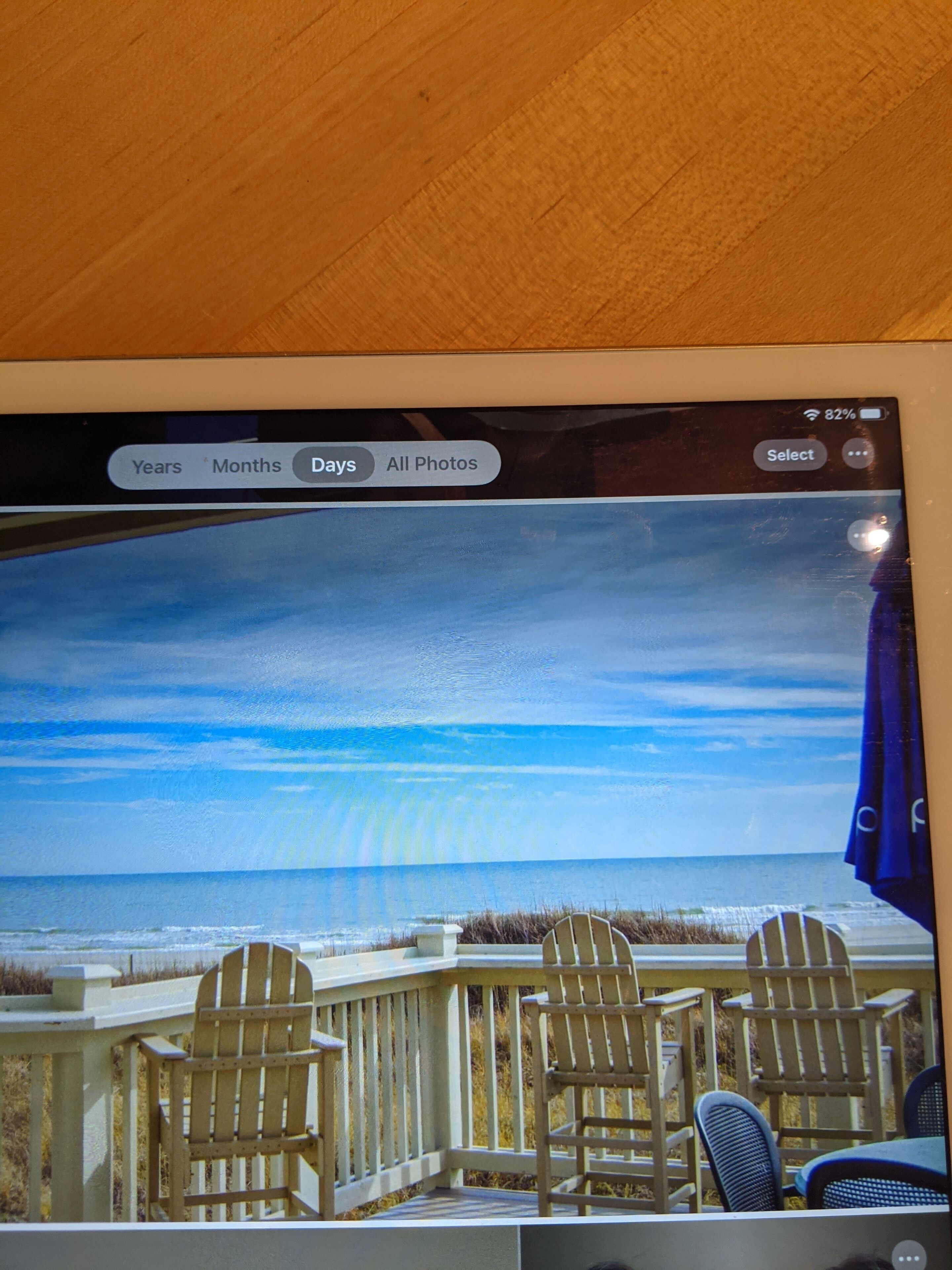 Interfaz de álbum de fotos con escena de playa, que incluye sillas y una mesa de madera, y un teléfono inteligente mostrando una foto con indicador de batería.