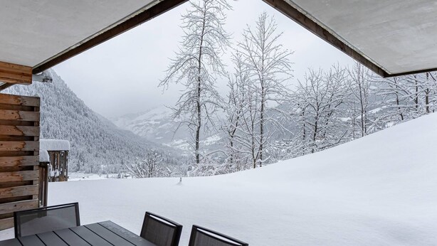 Un tavolo e sedie in legno in un ambiente esterno innevato, con vista su alberi e montagne ricoperte di neve.