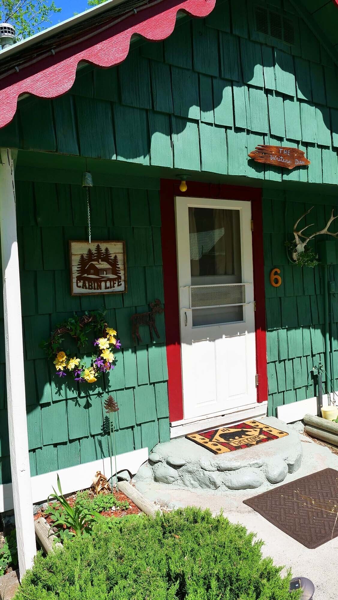 Une cabane à l’extérieur vert, avec une porte rouge et une pancarte indiquant « Vie à la cabane ».