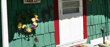 Une cabane à l’extérieur vert, avec une porte rouge et une pancarte indiquant « Vie à la cabane ».