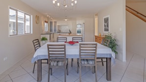 Een eethoek met een tafel voor vier personen, omringd door houten stoelen met gestreepte kussens. Een kroonluchter hangt aan het plafond, en er is een keuken in de achtergrond zichtbaar.