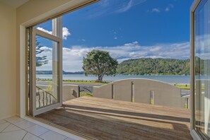 A room with a large glass door leading to a balcony with a view of a lake and a tree.