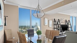 A modern living room with a glass dining table, woven chairs, a grey sofa, and a city view through large windows. - Triple en-suite unit with miles-long views of the Gulf | Beach Chairs Included (Panama City Beach)