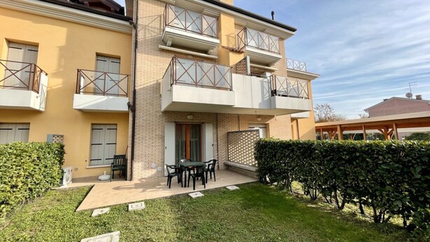 A two-story residential building with a balcony on the second floor, a small patio with a table and chairs, and a well-maintained garden.