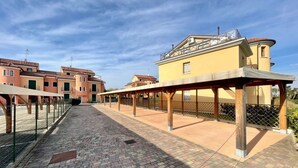 A covered outdoor walkway with a brick floor, leading to a yellow building with a balcony.